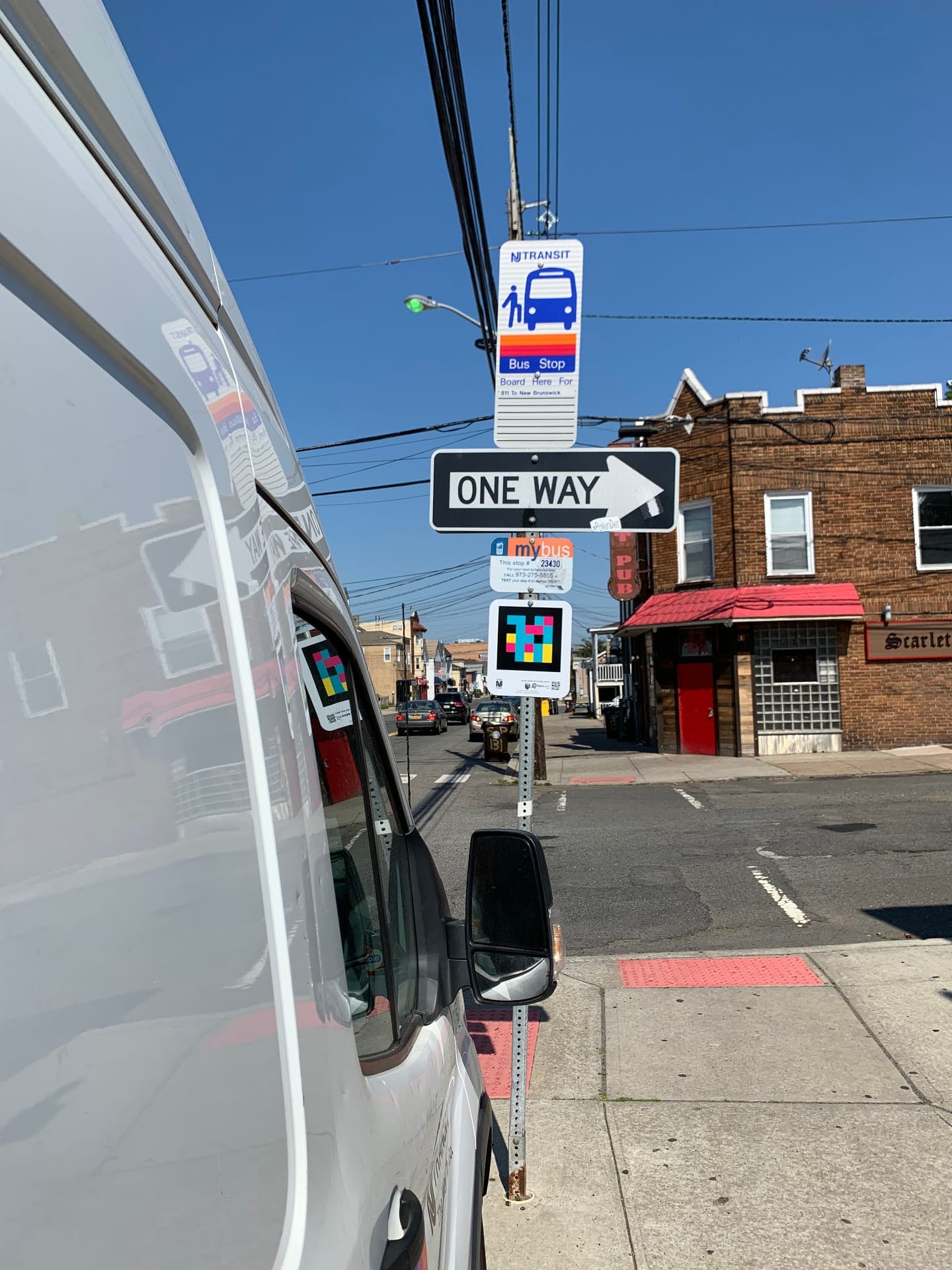 Corner with an NJ TRANSIT sign, One Way sign and NaviLens code under the mybus #23430 placard, in front of a pub in New Brunswick